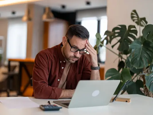 Verzweifelter Bauherr am Tisch mit Laptop, Taschenrechner und Papieren – steht sinnbildlich für finanzielle Belastung durch Bau-Insolvenz ohne passenden Schutz beim Hausbau
